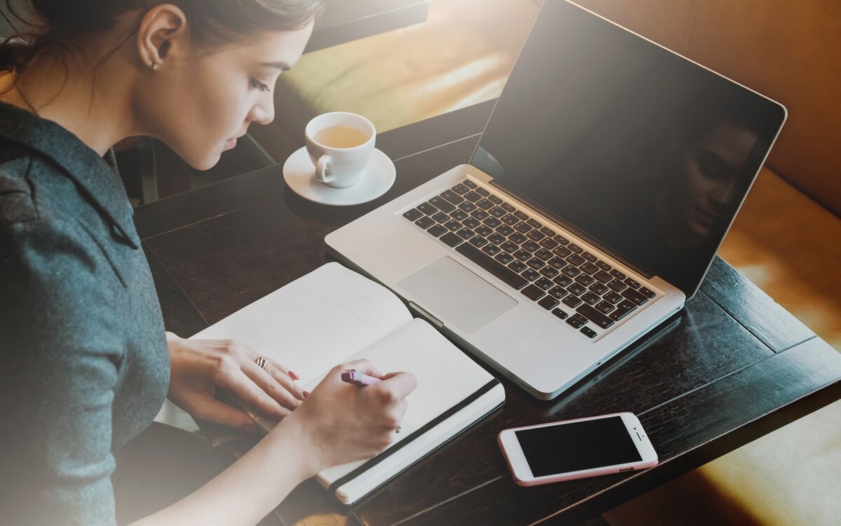 Woman writing in a notebook at a table with a laptop, smartphone, and cup of coffee.